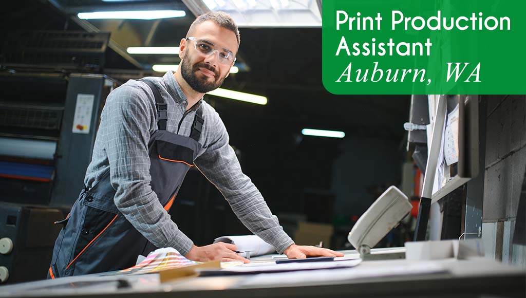 A man smiles and checks a print job for accuracy in a print production environment. White text over a green banner reads: 'Print Production Assistant in Auburn, WA.